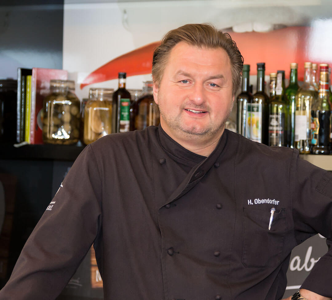 Herr Obendorfer steht mit einer Kochschürze in einer Küche und lacht in die Kamera.