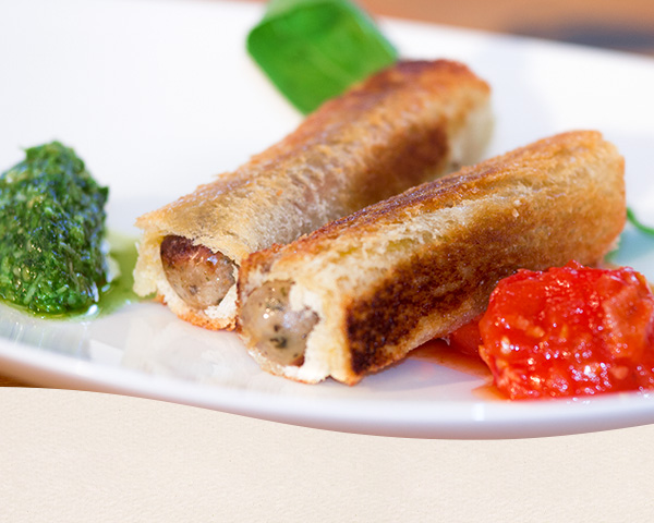 Nahaufnahme von Nürnberger Bratwürsten ummantelt von einem Tramezzini und gebraten. Dazu ein grünes Pesto und Tomatensoße.