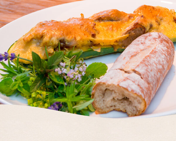 Nahaufnahme einer halben Zucchini gefüllt mit Nürnberger und mit Käse überbacken. Dazu Salat und Baguette.