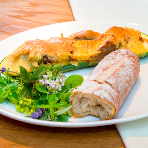Nahaufnahme einer halben Zucchini gefüllt mit Nürnberger und mit Käse überbacken. Dazu Salat und Baguette.